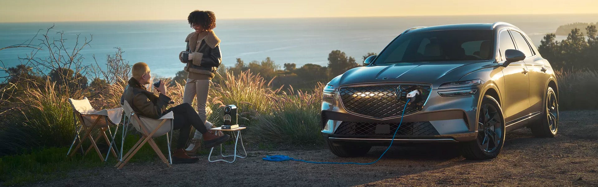 An all new Maluka Gray 2024 Genesis Electrified GV70 plugged in on a coastline with a couple enjoying some morning beverages. - Genesis of Baton Rouge, LA