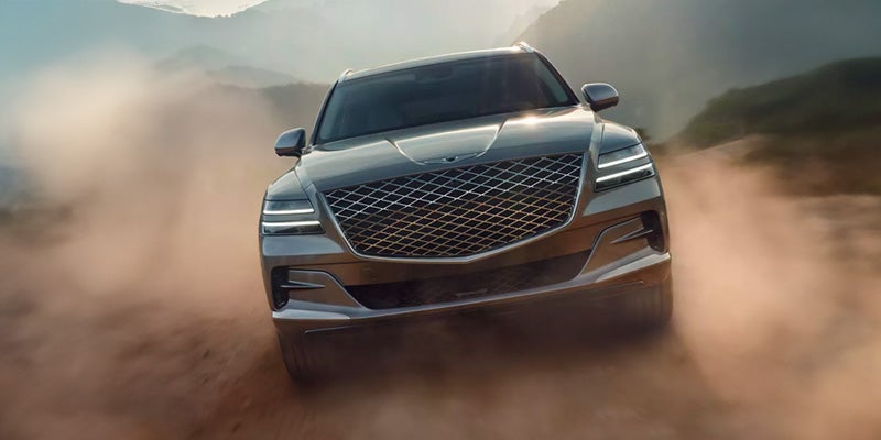 An all-new grey 2024 Genesis GV80 cruising a dirt road through forested hills. - Genesis of Baton Rouge, LA