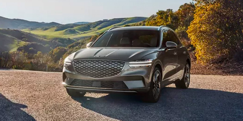 An all-new Gray 2024 Genesis GV70 parked on top of a gravel driveway surrounded by rolling green hills. - Genesis of Baton Rouge, LA