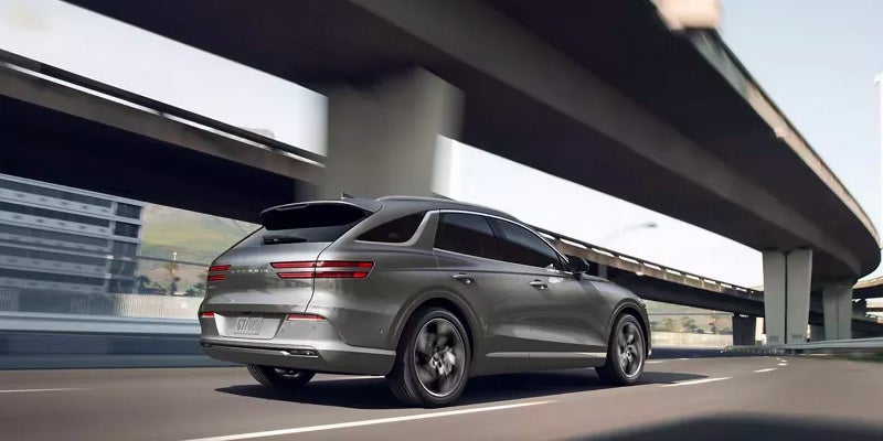An all-new Silver 2024 Genesis Electrified GV70 driving on a downtown freeway under an overpass. - Genesis of Baton Rouge, LA