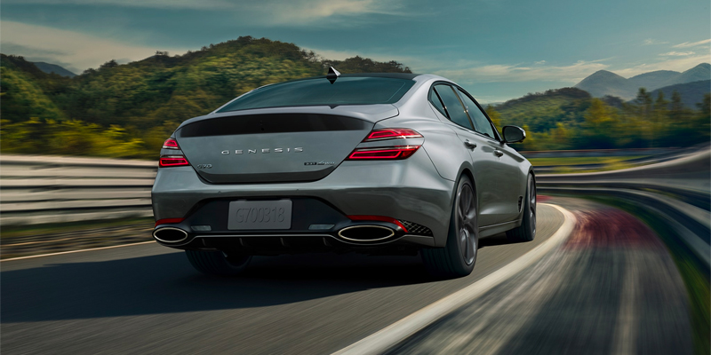 A Silver Genesis sedan driving down on off-ramp among forested hills. - Genesis of Baton Rouge, LA