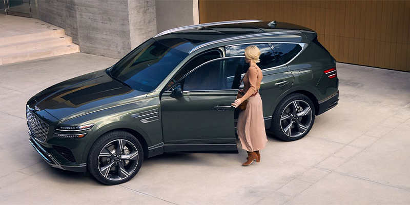 A woman opening the driver door of a dark green Genesis GV80 luxury midsize SUV parked in a driveway.